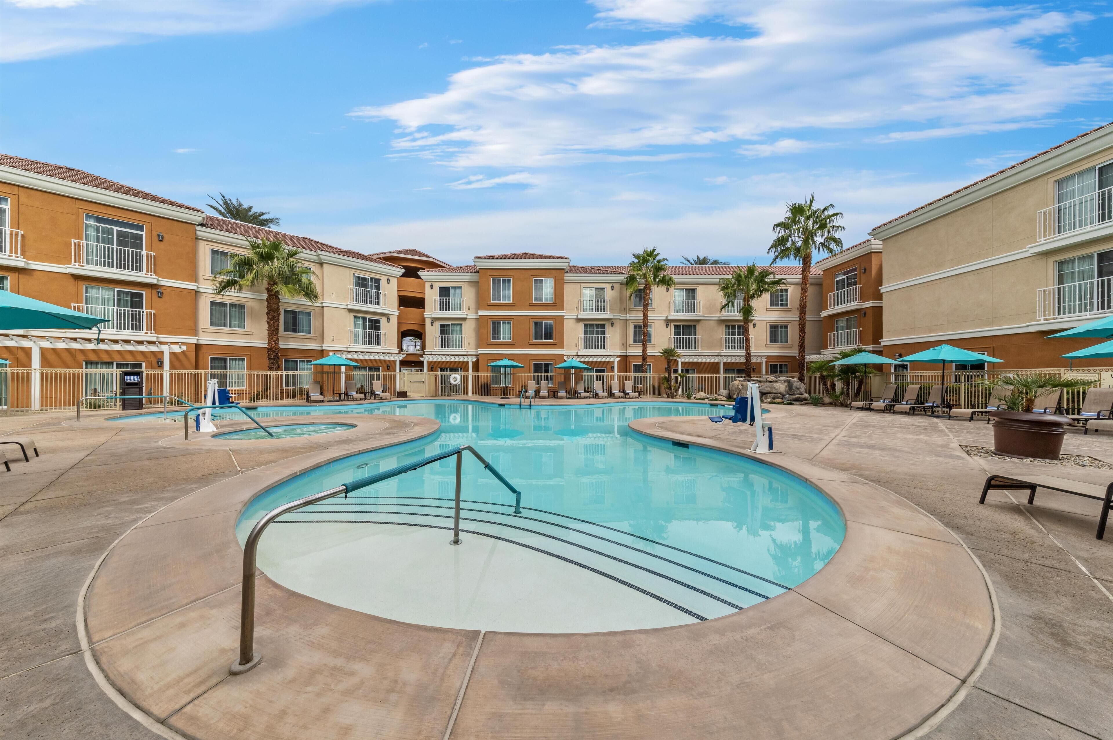 A pool area with multiple lounge chairs and umbrellas, surrounded by multi-story buildings with balconies.