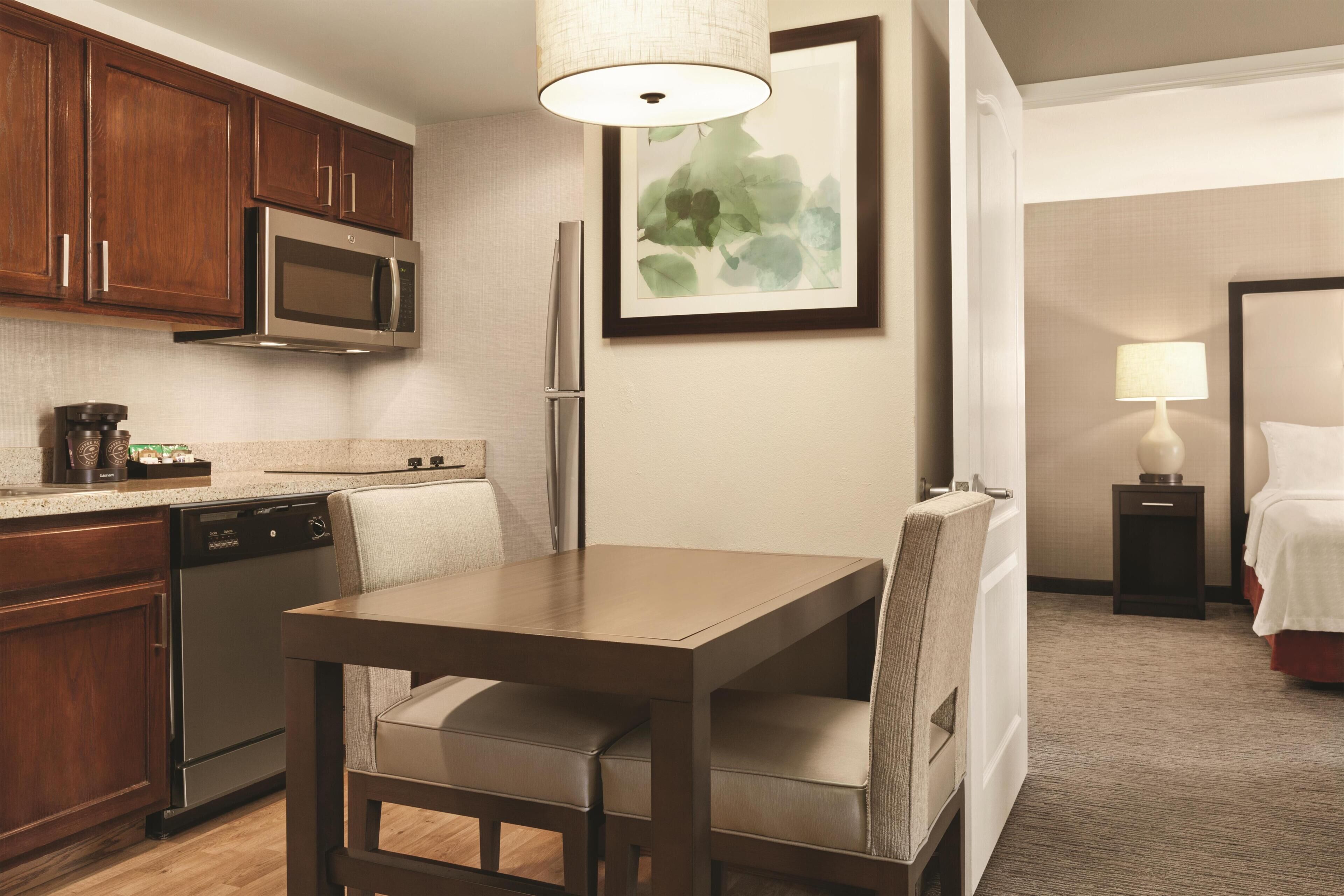 Quarto de hotel com cozinha, mesa de jantar com cadeiras, cama, mesa de cabeceira com abajur e uma obra de arte emoldurada na parede.