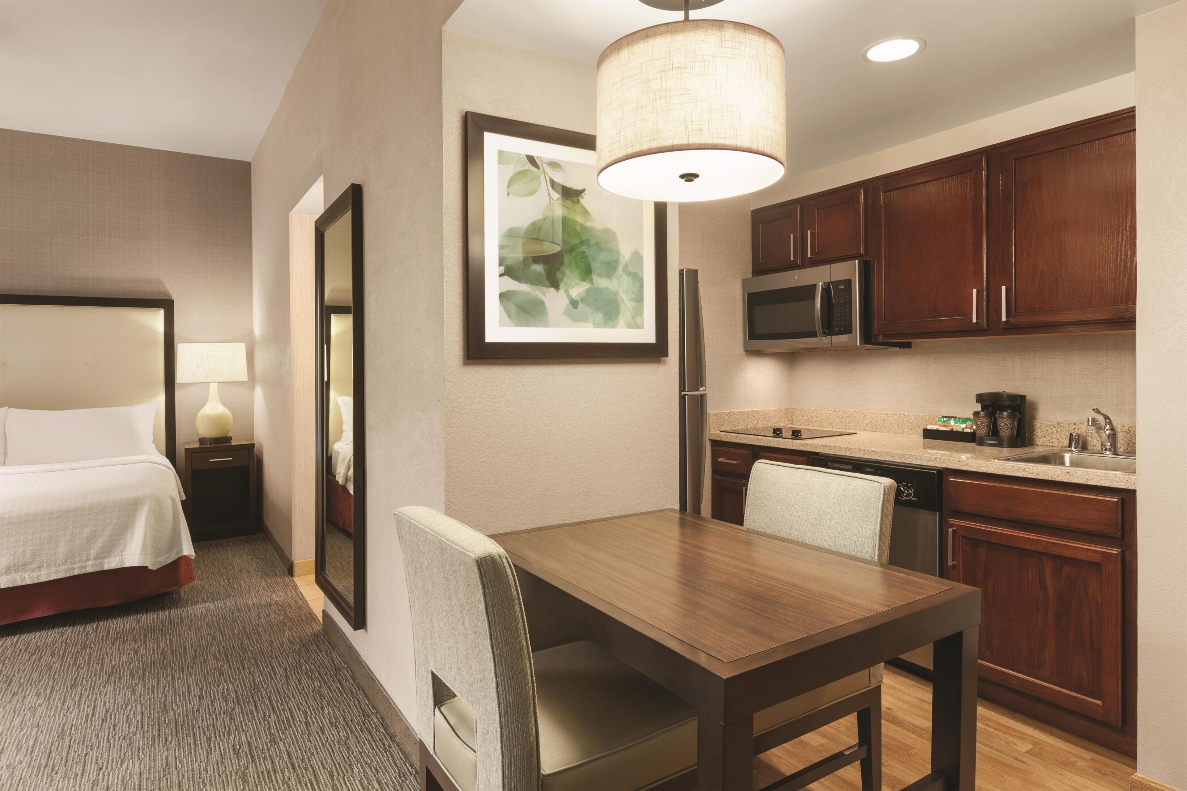 A hotel room with a bed, a dining table, a chair, a kitchen area with a microwave, and a refrigerator.