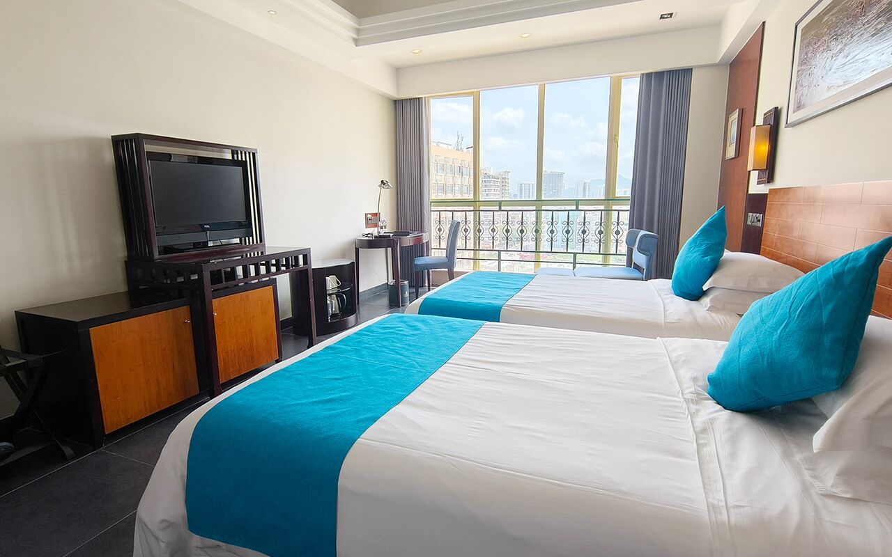 A hotel room with two beds, a TV, a desk, and a balcony with a city view.