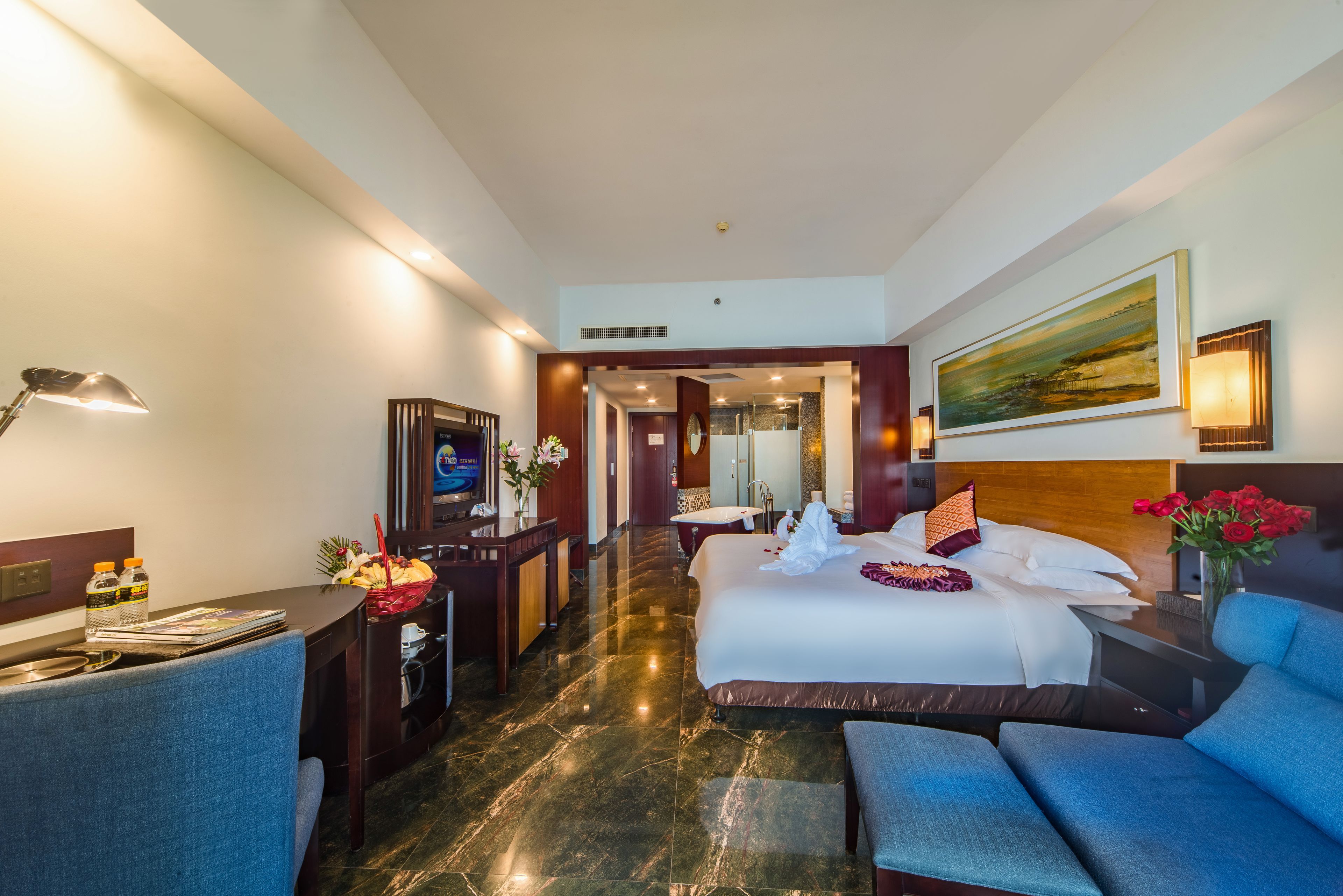 A hotel room with a large bed, a desk with a lamp, a chair, a painting on the wall, and a view of the hallway.
