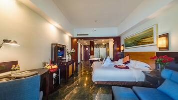 A hotel room with a large bed, a desk with a lamp, a chair, a painting on the wall, and a view of the hallway.