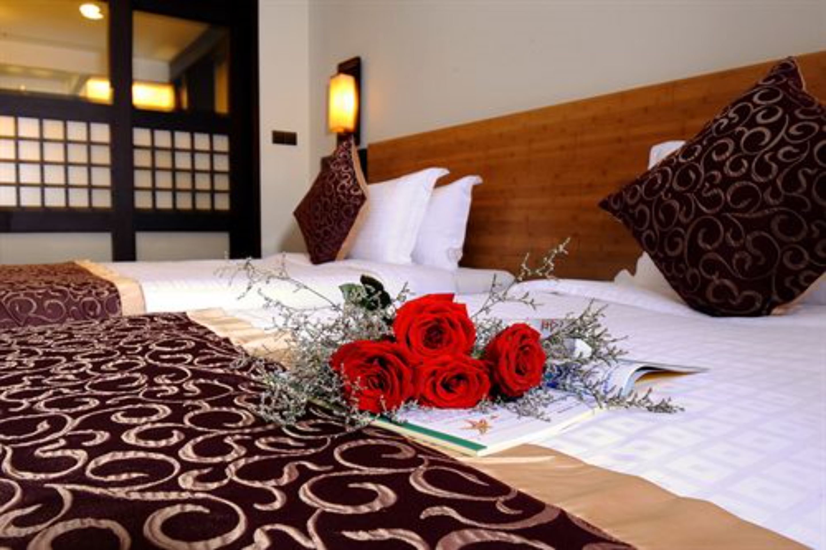 A hotel room with a bed, a bedside table with a lamp, and a decorative arrangement of red roses and greenery.