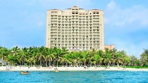 Ein hohes Hotel mit mehreren Etagen, ein Strand mit Palmen und ein klarer, blauer Himmel.