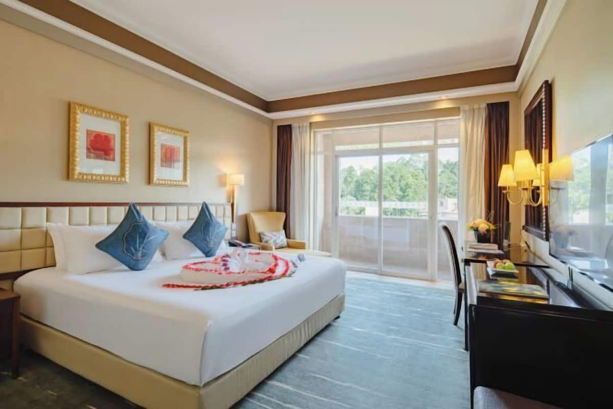 A hotel room with a large bed, a desk with a chair, a TV, and a balcony with curtains.