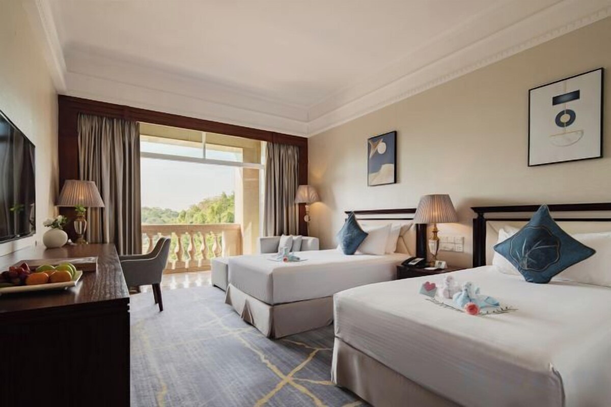 A hotel room with two beds, a desk with a chair, a dining table with fruits, and a balcony with curtains.
