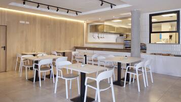 A modern dining area with white tables and chairs, a wooden wall, and a kitchen in the background.