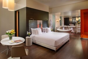 Una moderna camera d'hotel con un letto grande, un'area salotto con un tavolo e sedie, un piccolo tavolino con un vaso di fiori e un bagno con vasca e lavabo.