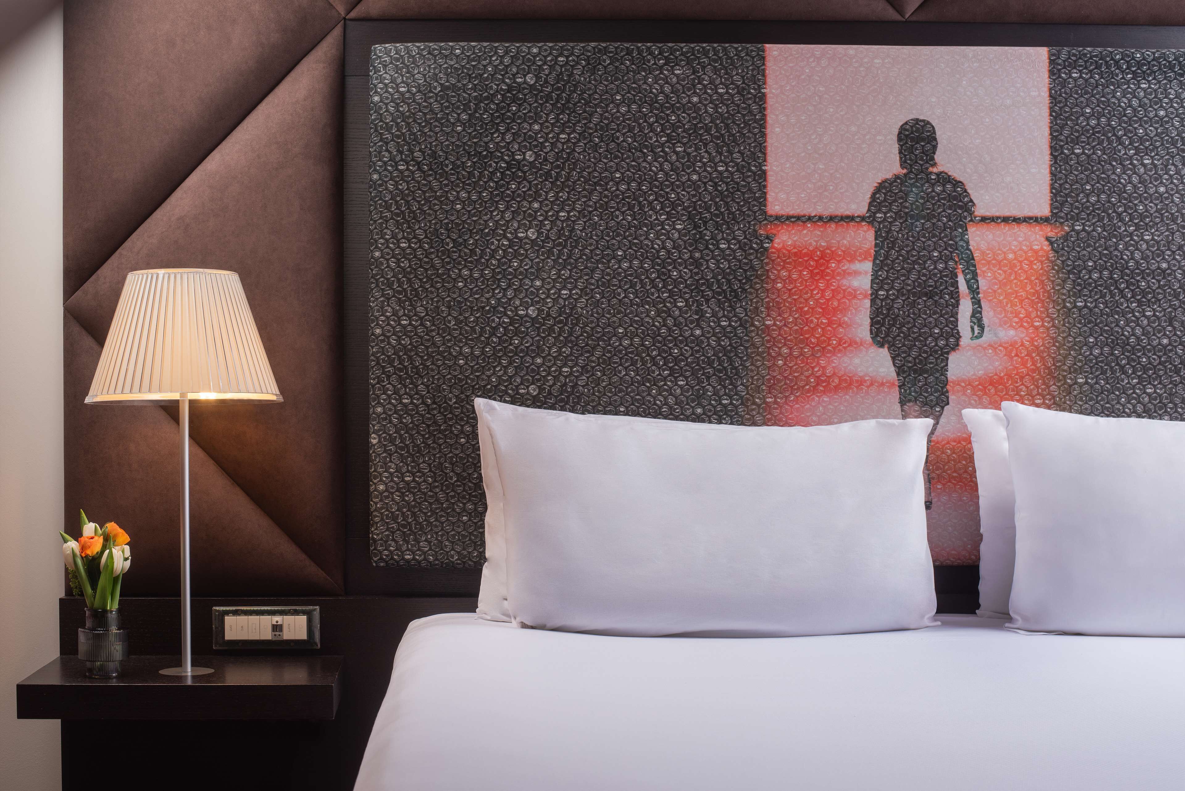 Une chambre d’hôtel avec un lit, des oreillers blancs, une table de chevet avec une lampe et une œuvre d’art accrochée au mur.