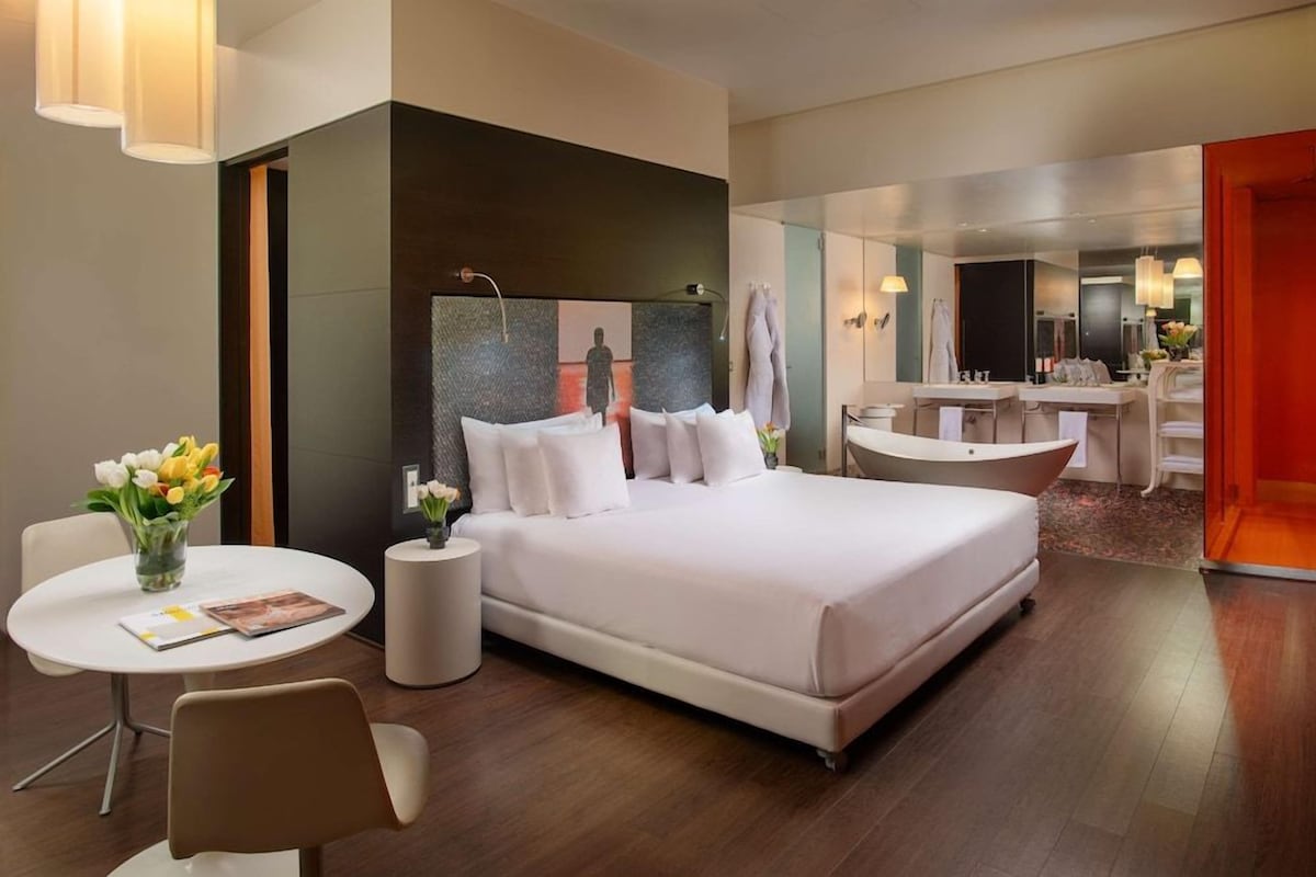 Una habitación de hotel moderna con una cama grande, una zona de estar con una mesa y sillas, un baño con bañera y un espejo.