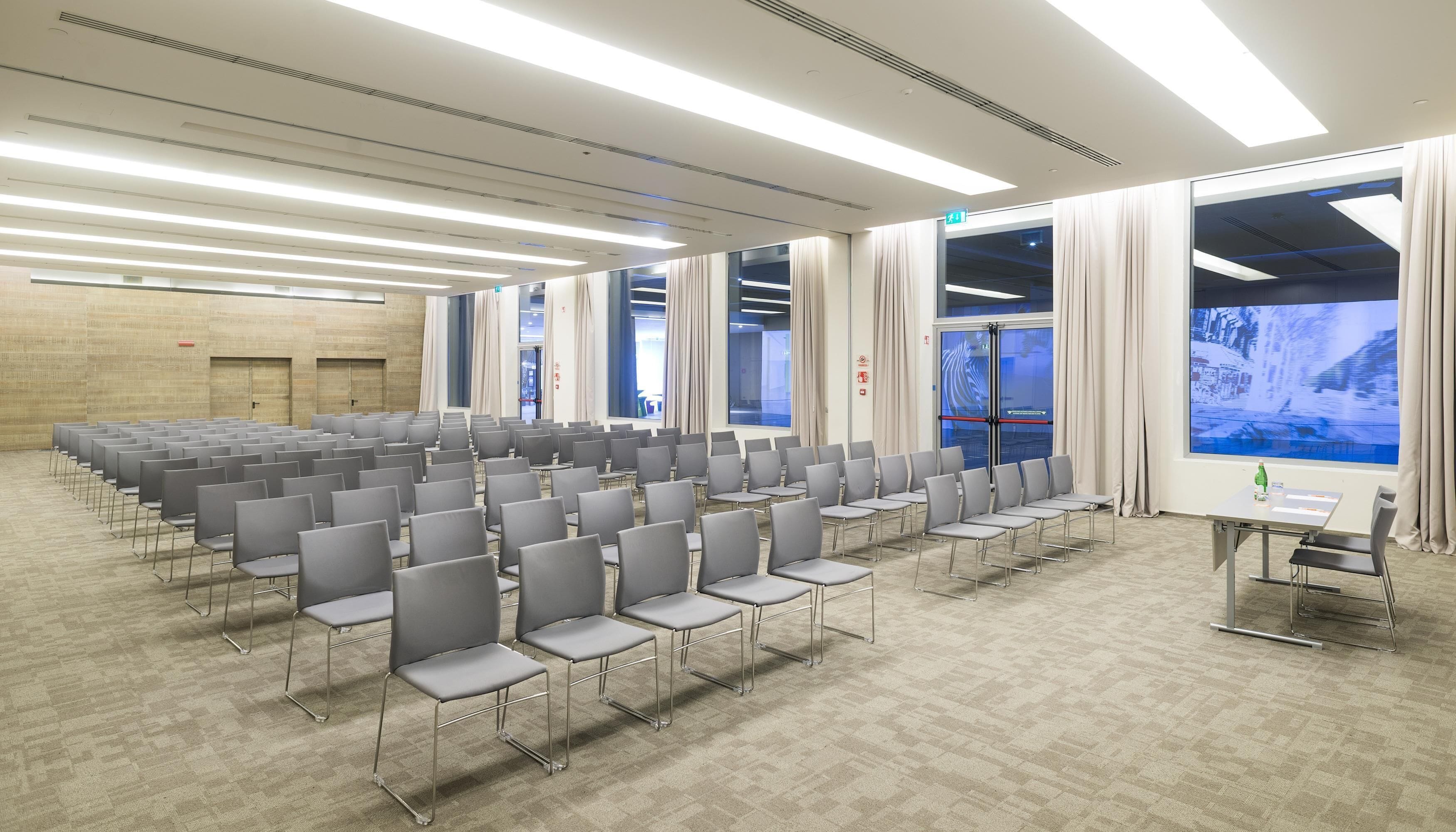 A conference room with rows of chairs, large windows with curtains, and a projection screen.