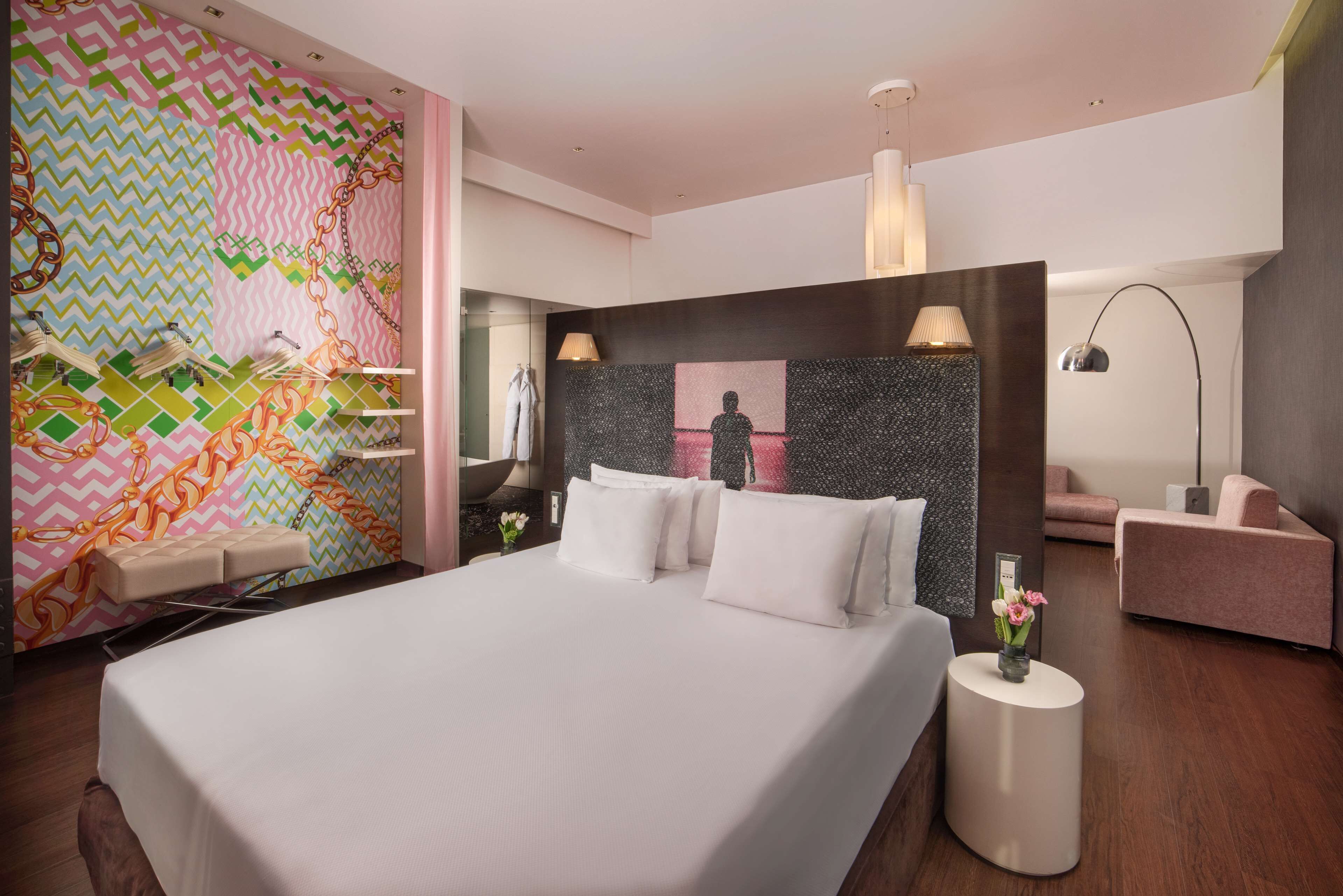 A modern hotel room with a large bed, a sitting area with a chair, a small table with a vase, and a mirror.
