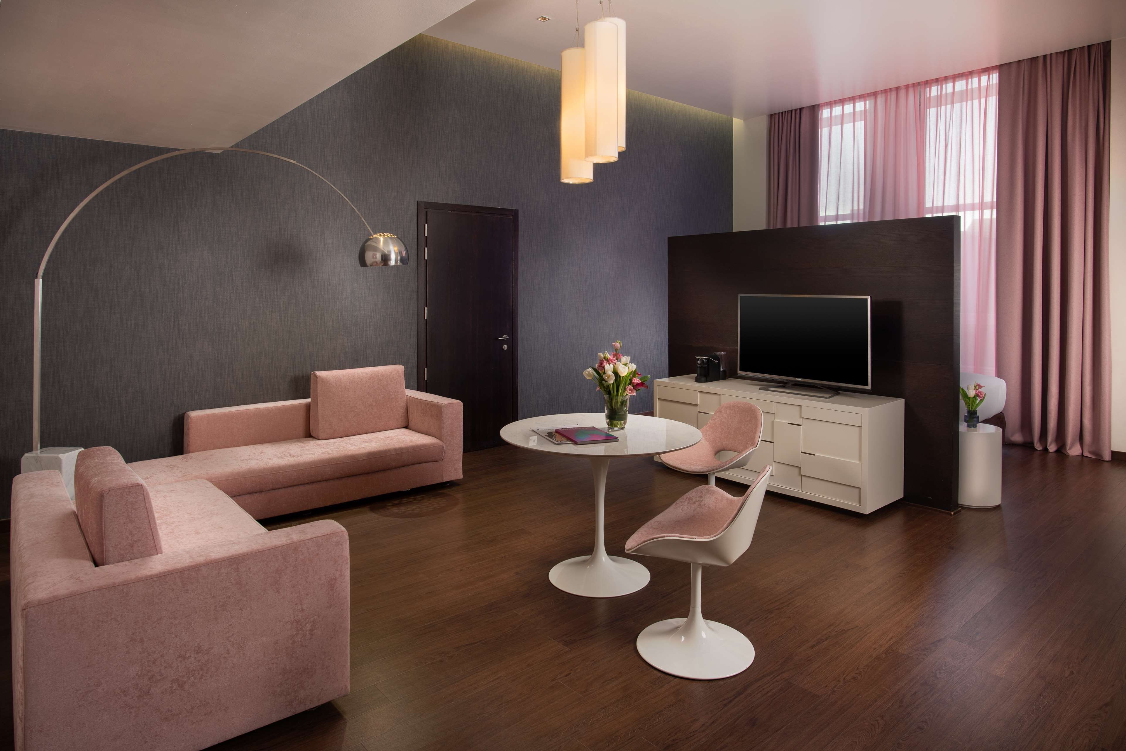A modern living room with a sofa, two chairs, a round table, a TV, and a large window with curtains.