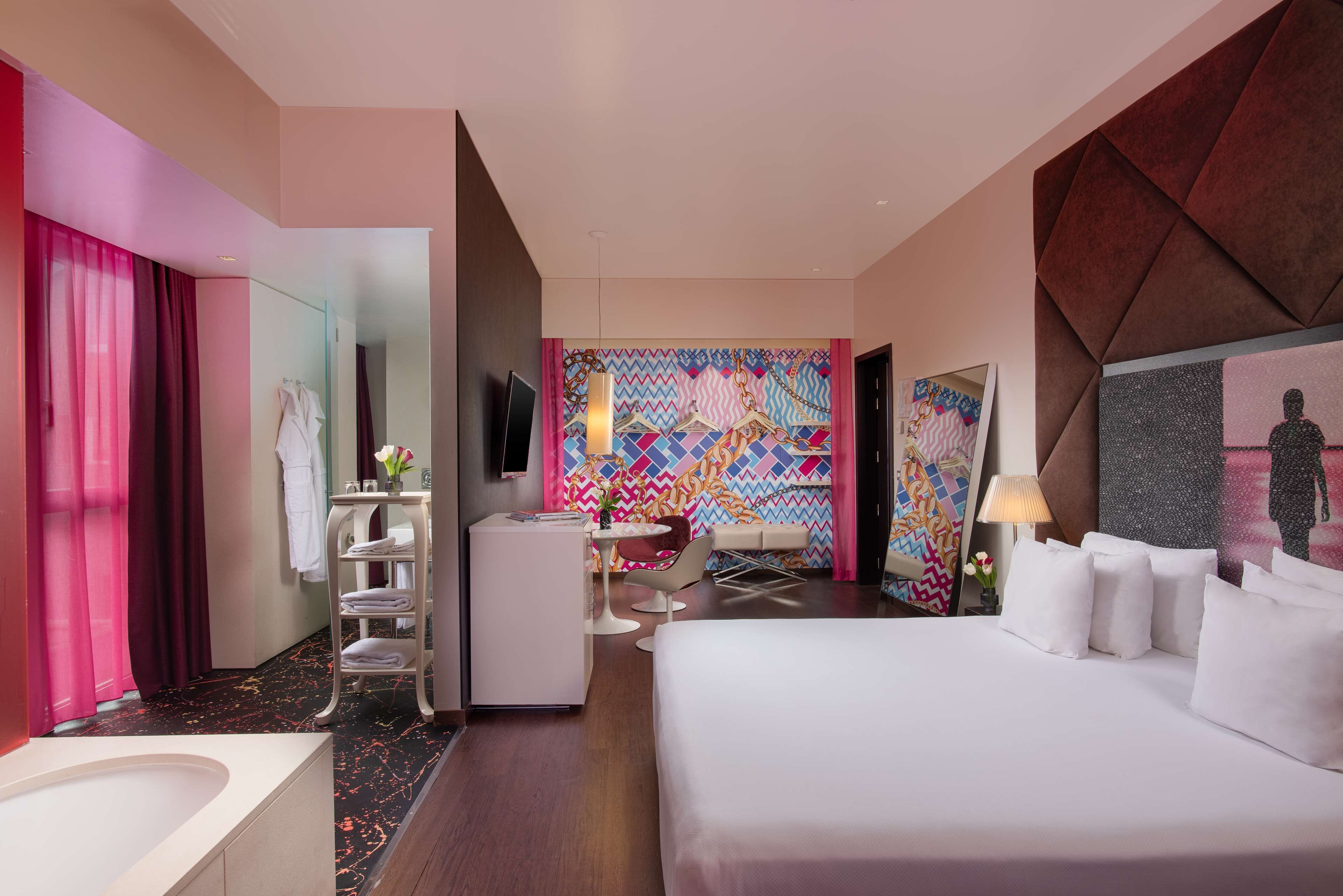 A modern hotel room with a large bed, a desk with a chair, a vanity with a mirror, and a colorful wall mural.