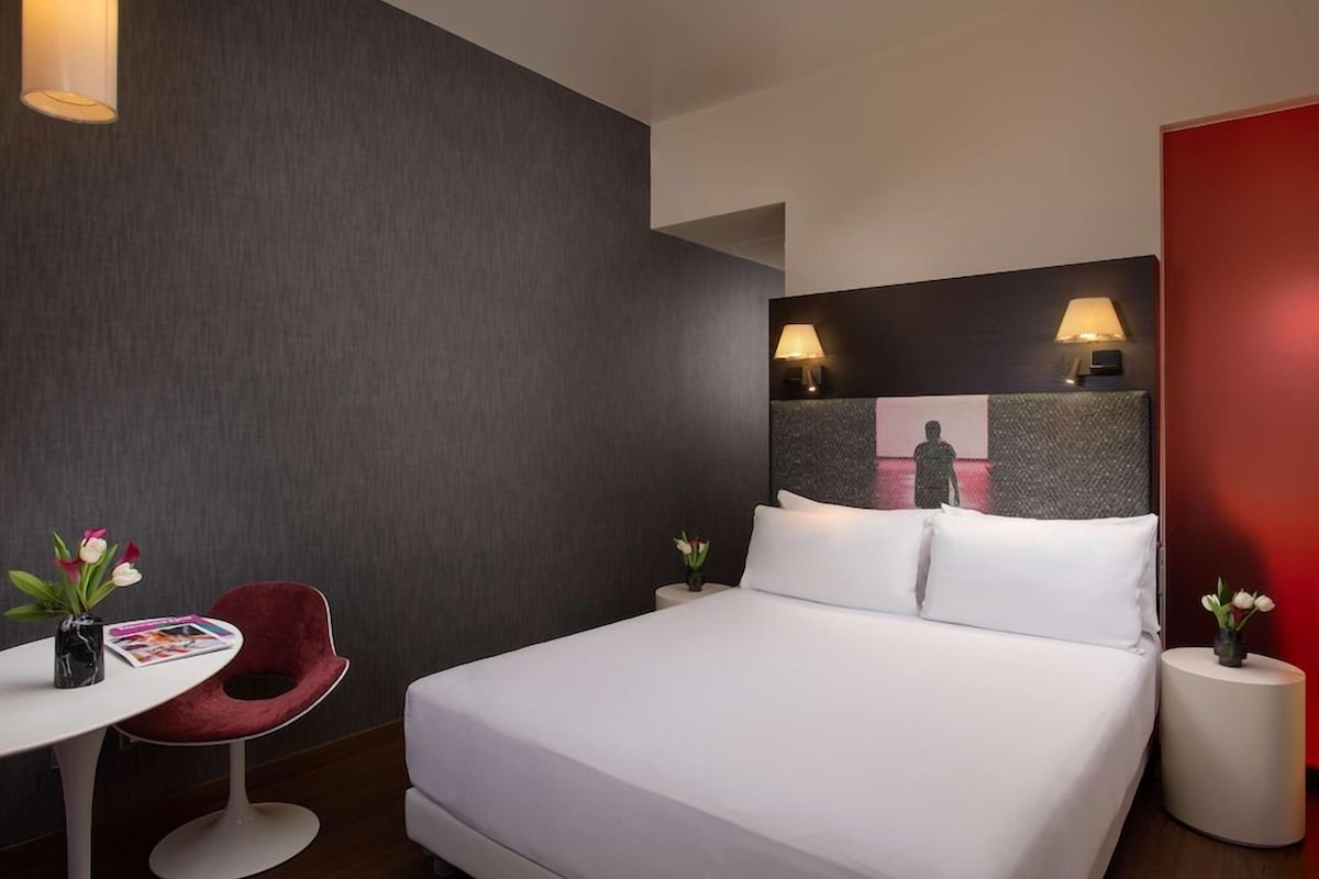 Una habitación de hotel moderna con una cama grande, dos mesitas de noche, una pared de acento roja y una pequeña mesa con una silla y flores.