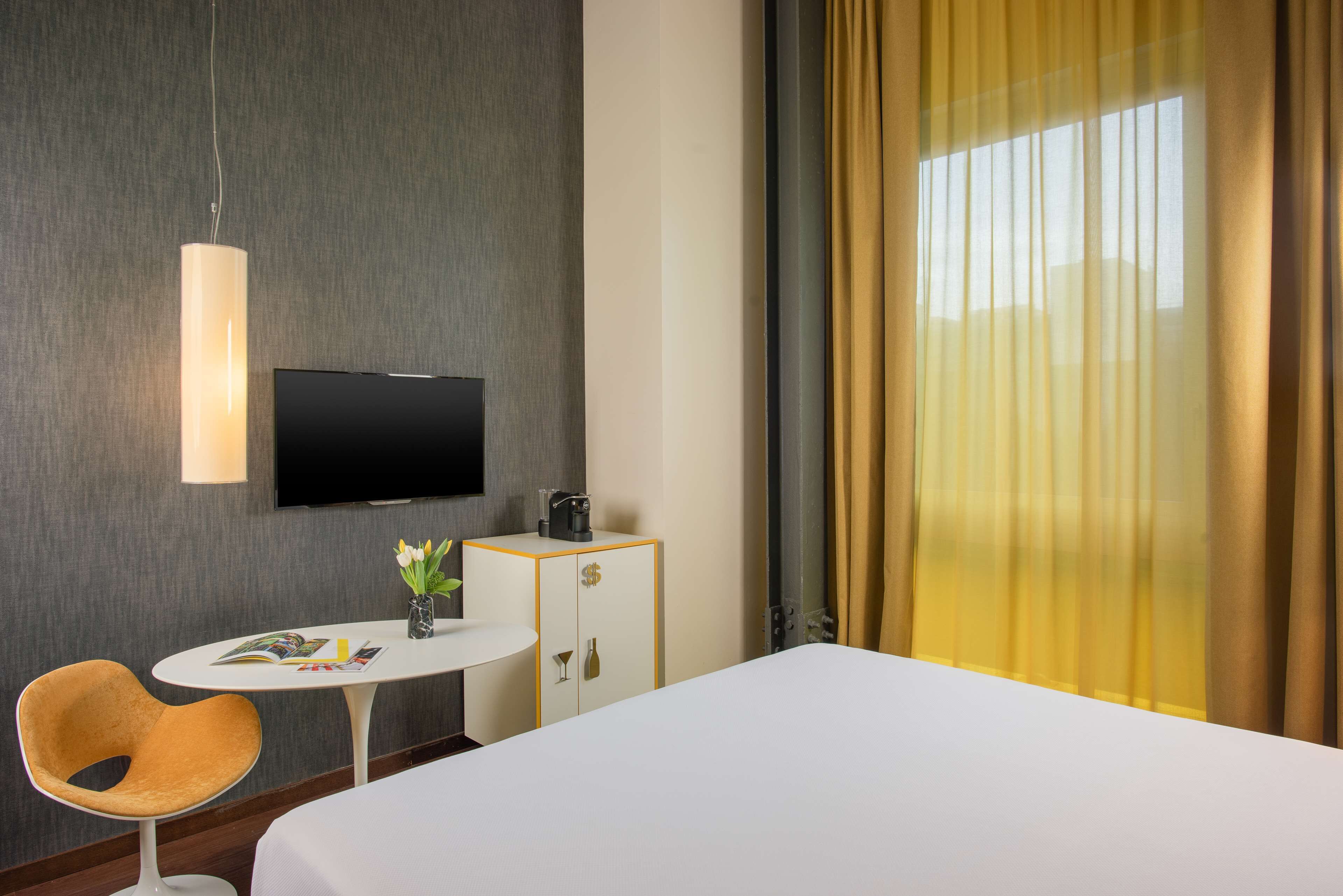 Een hotelkamer met een bed, een aan de muur bevestigde televisie, een tafeltje met een tijdschrift, een stoel en een kast met een koffieapparaat.