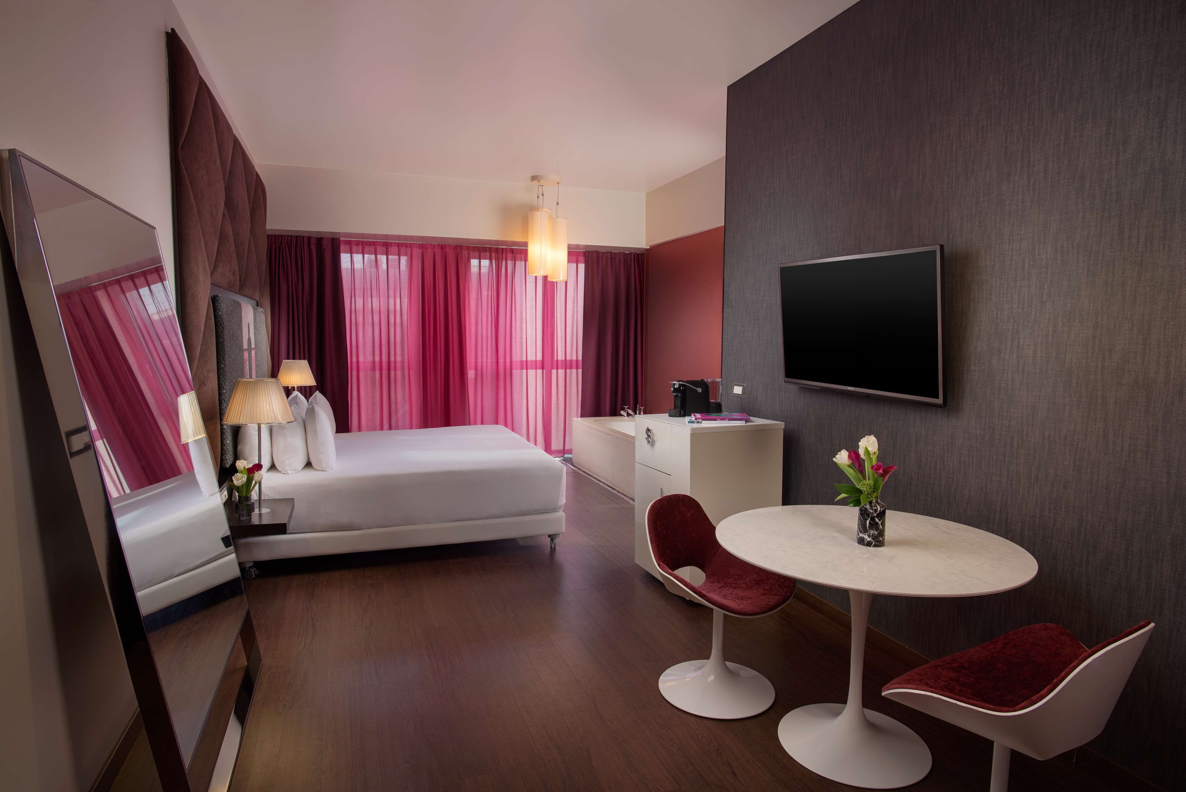 Una habitación de hotel moderna con cama, mesita de noche, lámpara y una pequeña mesa redonda con dos sillas.