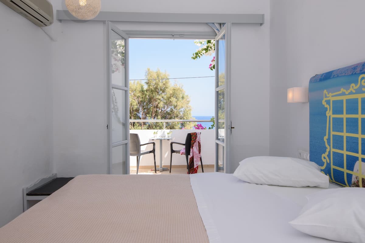 Un dormitorio con cama, una silla y una mesa. Hay un balcón con vista a árboles y al cielo.