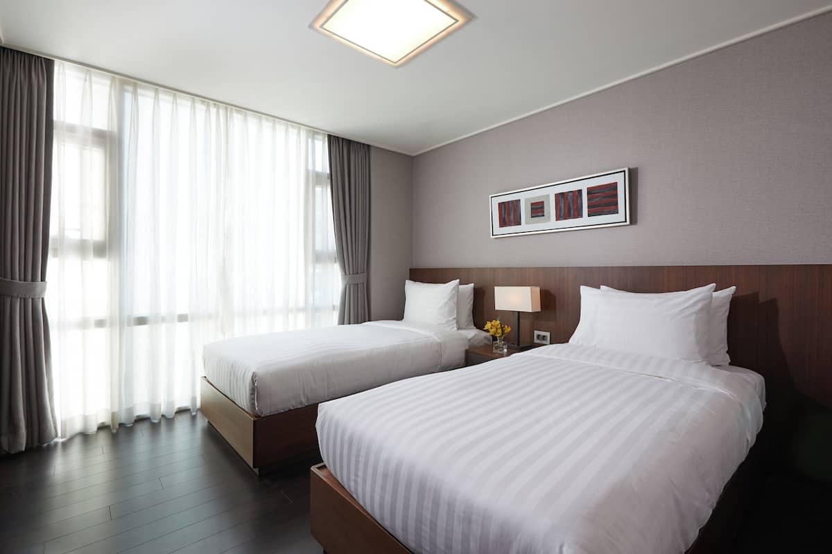 Habitación de hotel con dos camas, un ventanal con cortinas, un cuadro en la pared y una mesita de noche con lámpara.
