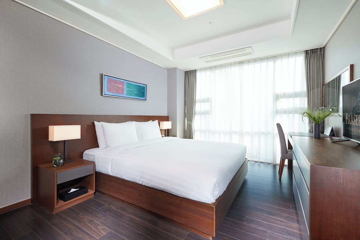 Habitación de hotel con una cama grande, un escritorio con televisor, una mesita de noche y una ventana con cortinas.