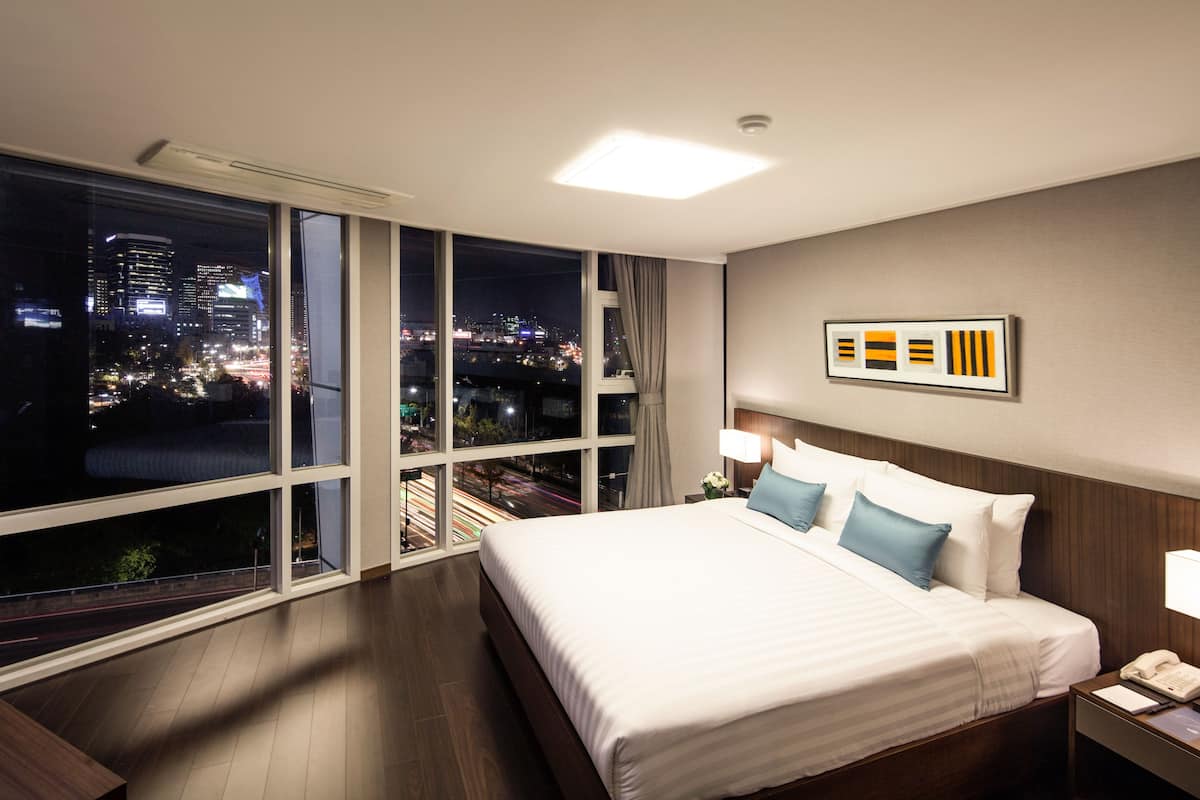Un cuarto de hotel con un amplio ventanal que ofrece vistas a la ciudad, una cama bien tendida, una mesita de noche con lámpara y un cuadro enmarcado en la pared.