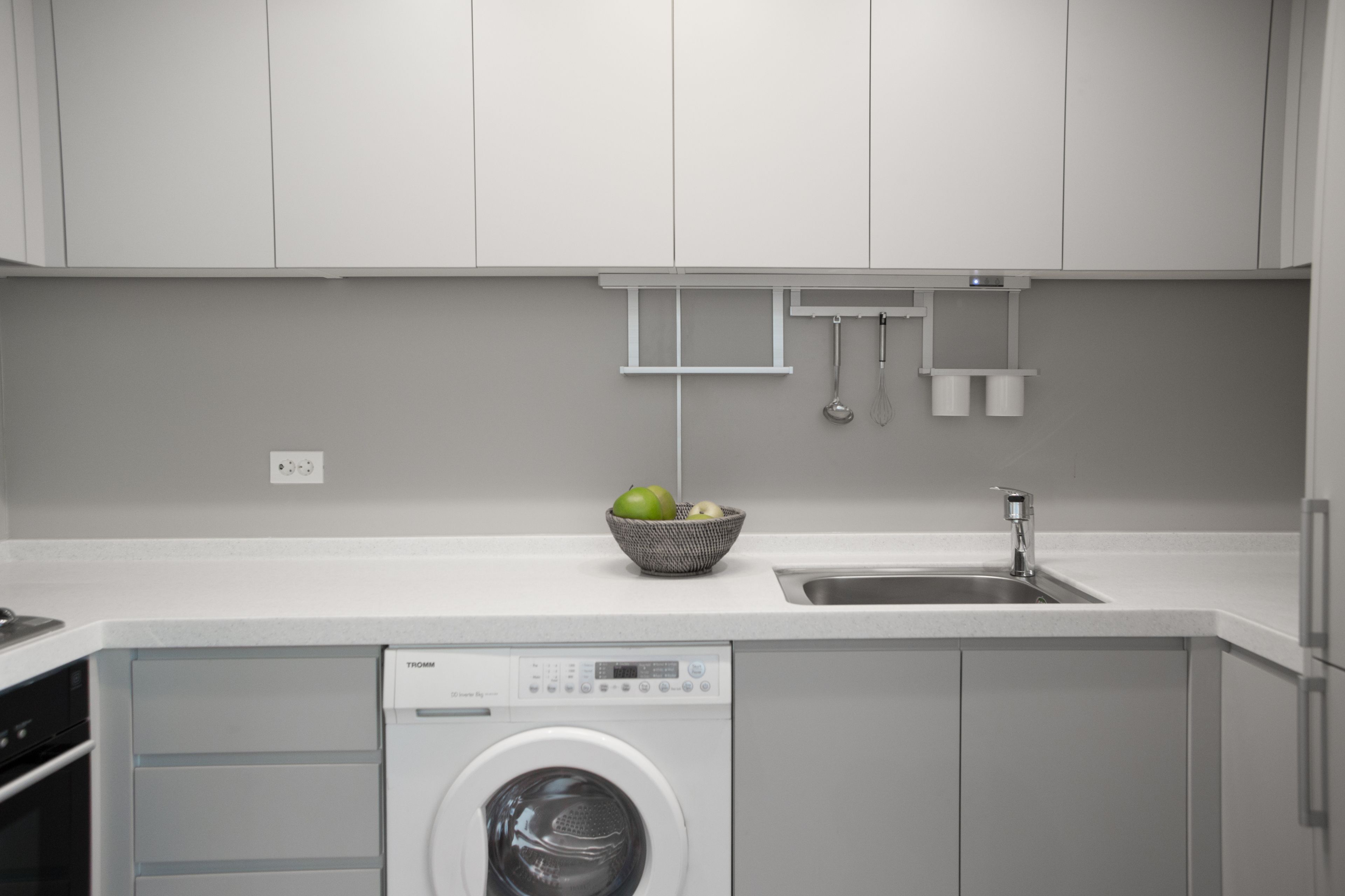 A modern kitchen with white cabinetry, a built-in washing machine, a sink, and a bowl of fruit on the countertop.