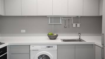 A modern kitchen with white cabinetry, a built-in washing machine, a sink, and a bowl of fruit on the countertop.