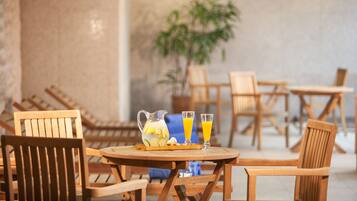 A wooden table with a pitcher and two glasses of orange juice, surrounded by wooden chairs in a room with a plant and a concrete wall.