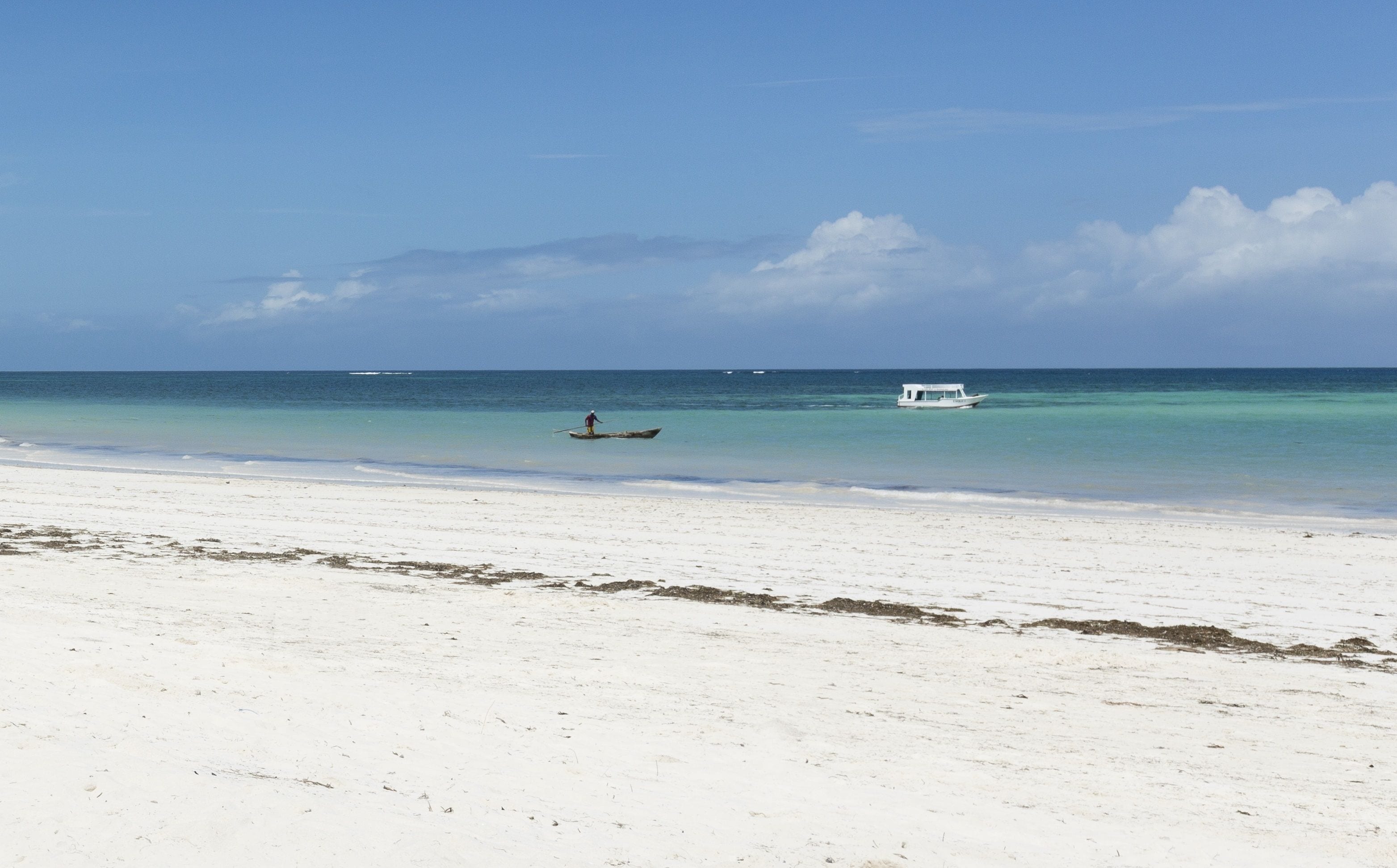 Une plage de sable blanc bordée d’eaux turquoise et calmes. On y voit une personne qui rame sur un petit bateau, et un bateau plus grand, ancré au loin.
