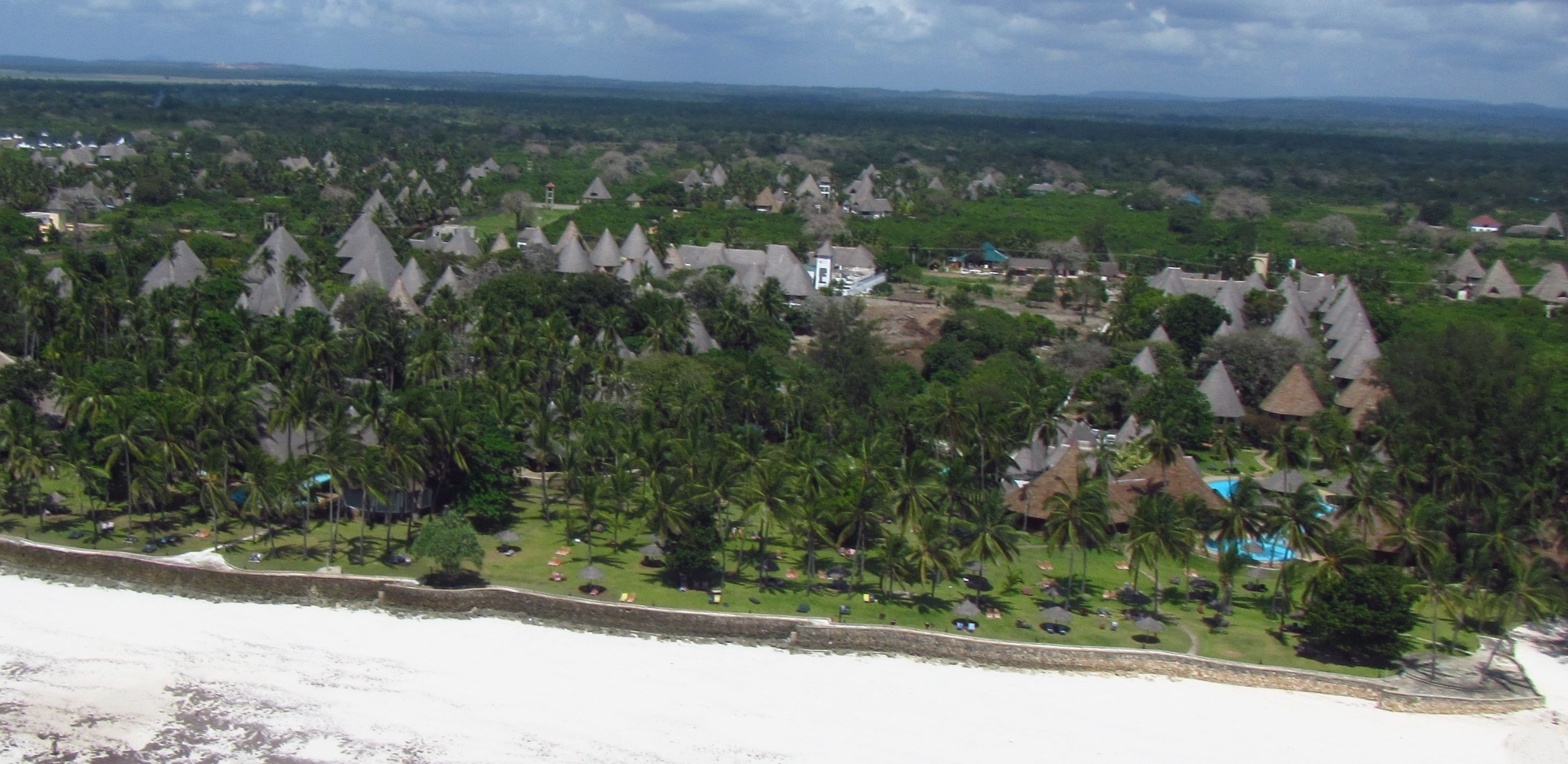 Un resort con edificios de techos cónicos, palmeras y una playa.