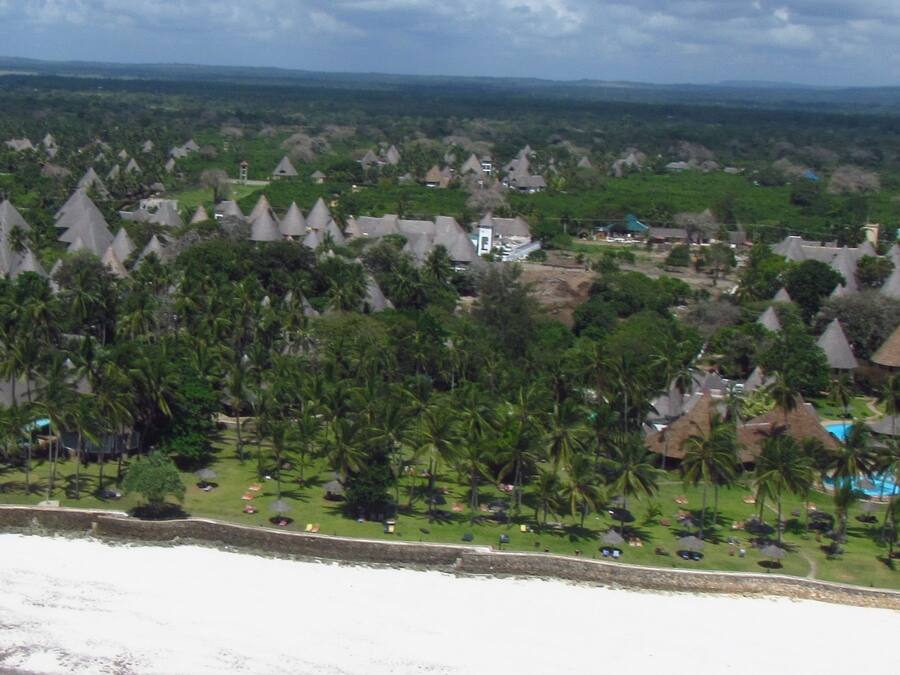 Un resort con edificios de techos cónicos, palmeras y una playa.