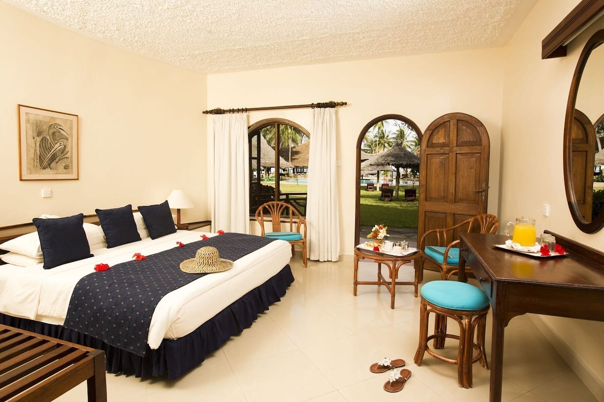 Quarto de hotel com cama, escrivaninha e cadeira, mesa de jantar e cadeira, além de vista para uma piscina e palmeiras.