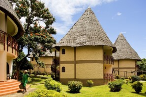 A traditional building with a thatched roof, a staircase leading to a balcony, and a well-maintained garden with various shrubs and a tree with orange flowers. - Neptune Paradise Beach Resort & Spa (Diani Beach)