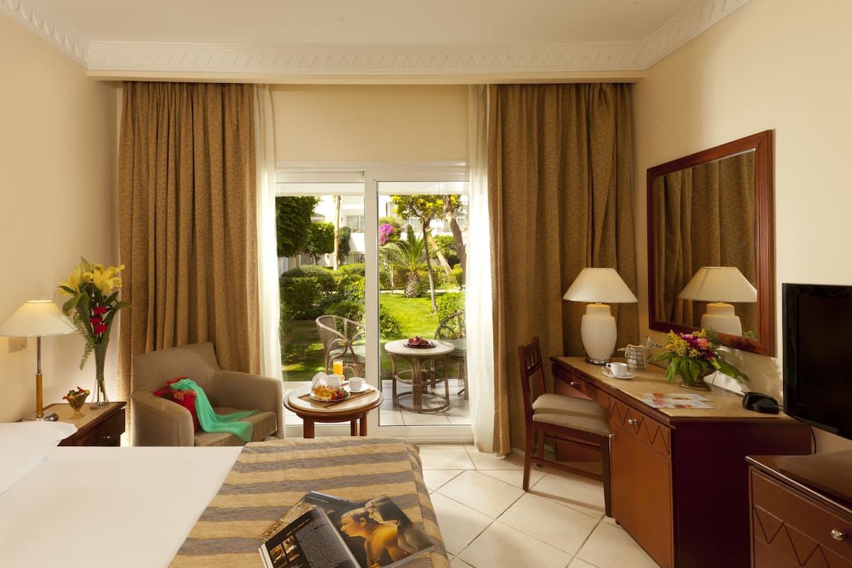 A hotel room with a bed, a desk, a chair, a small table with a fruit plate, a lamp, and a view of a garden through a glass door.