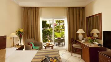 Ein Hotelzimmer mit Bett, Schreibtisch, Stuhl, einem kleinen Tisch mit Obstschale, einer Lampe und Blick auf einen Garten durch eine Glastür.