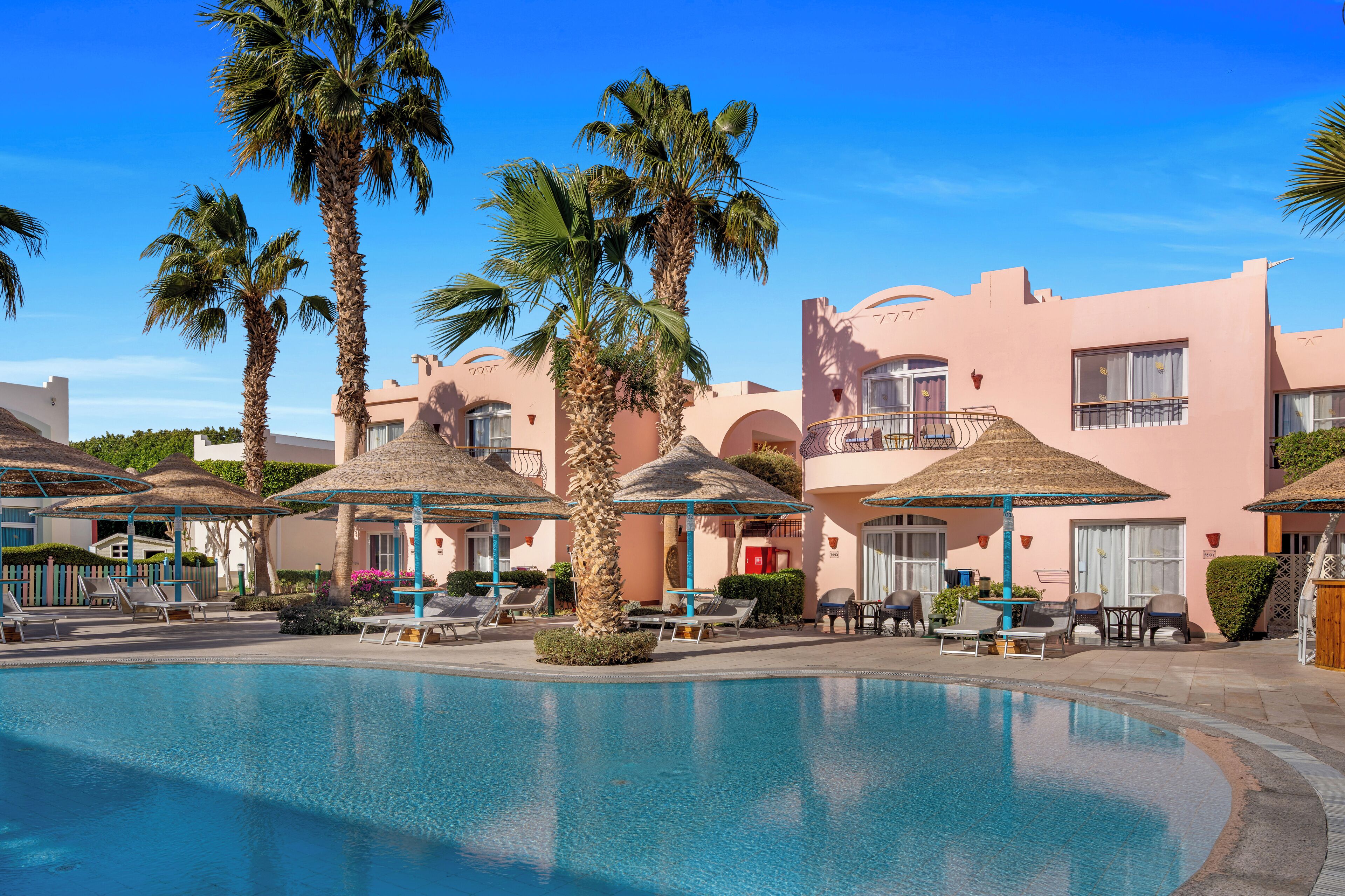 A pool area with lounge chairs and umbrellas, surrounded by palm trees and a pink building with balconies.