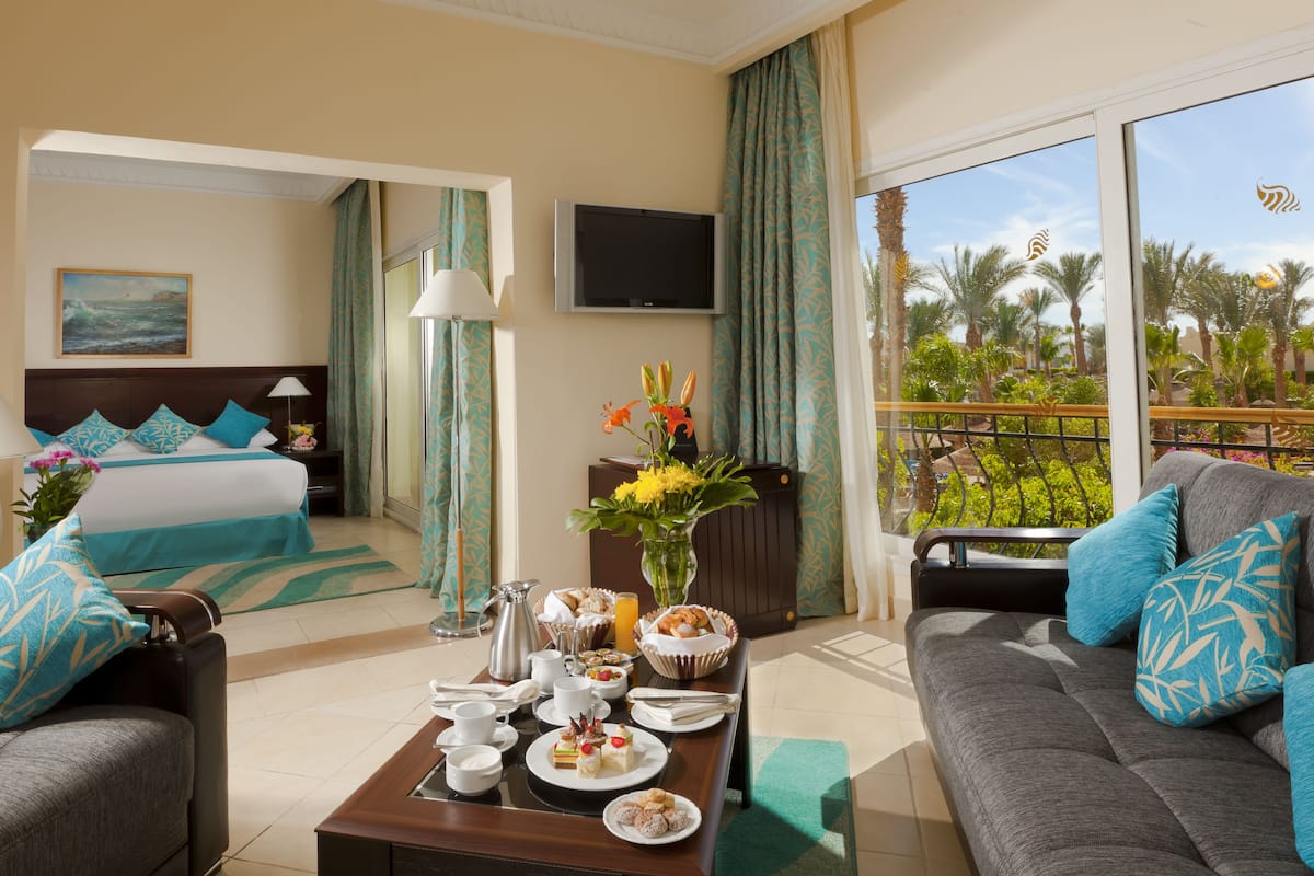 A hotel room with a bed, a sofa, a coffee table with breakfast items, a TV, and a balcony with a view of palm trees.
