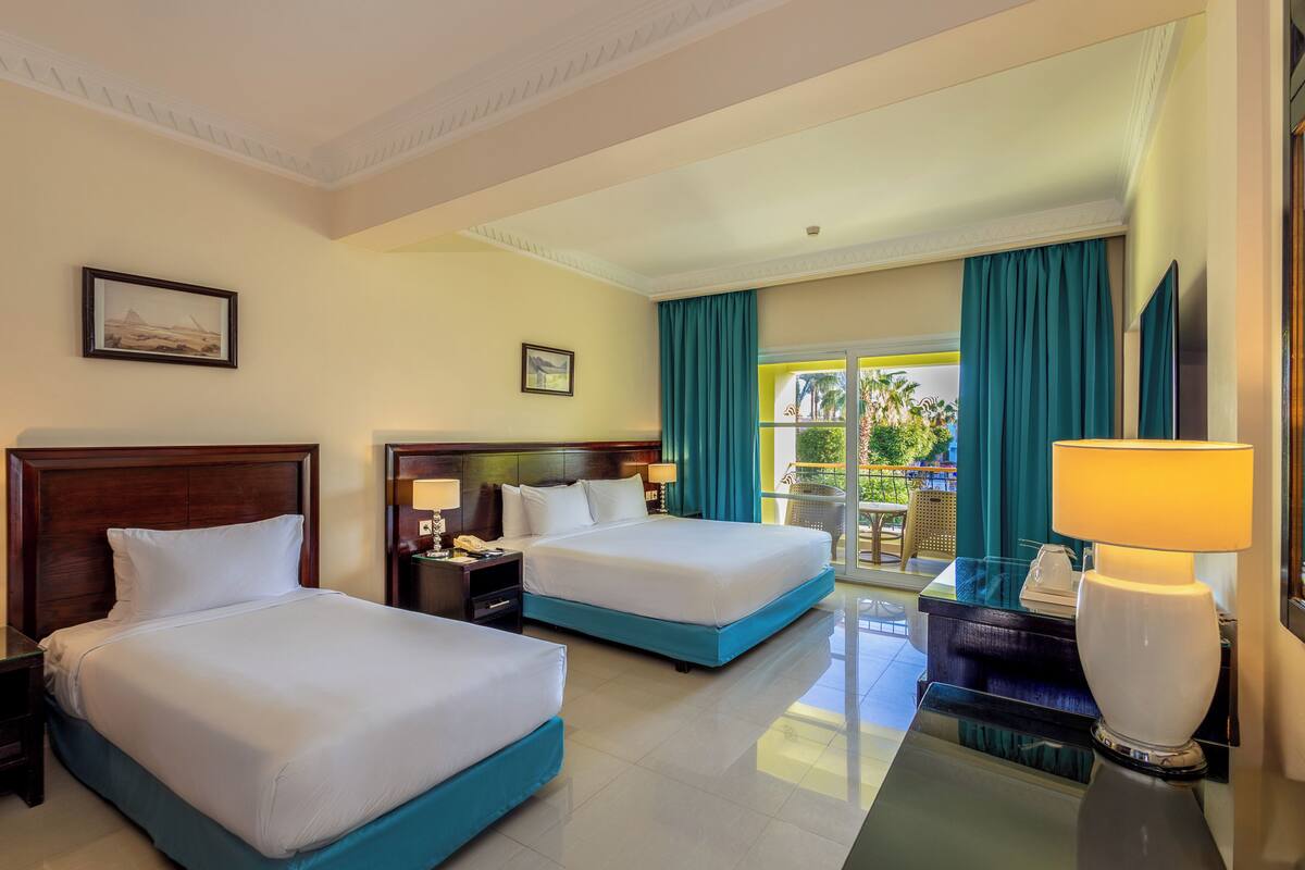 A hotel room with two beds, a nightstand with a lamp, a desk with a lamp, and a view of the outdoors.