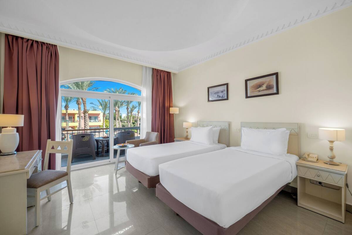 A hotel room with two beds, a desk, a chair, a table, and a balcony with a view of palm trees and buildings.