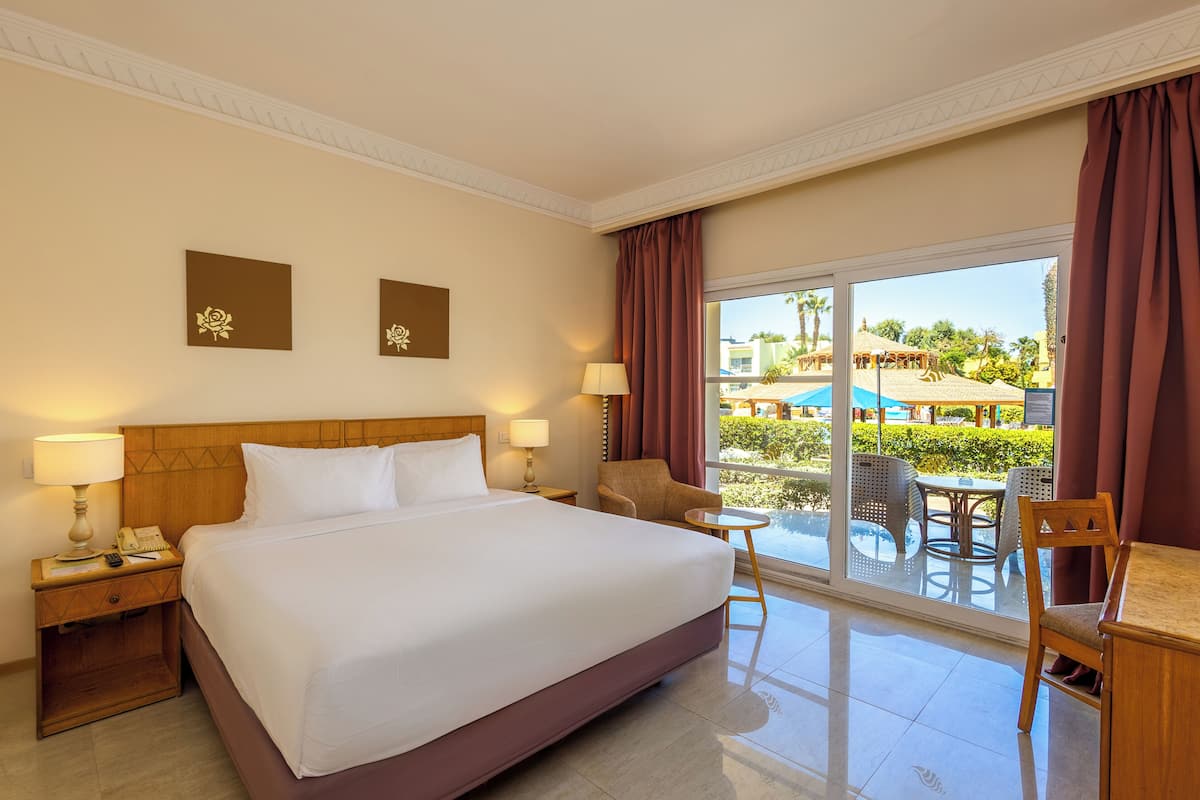 A hotel room with a large bed, two bedside tables with lamps, a chair, a small table, and a view of a pool and outdoor area.