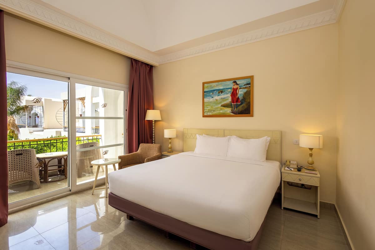 A hotel room with a large bed, two bedside tables, a chair, a balcony with a view, and a painting on the wall.