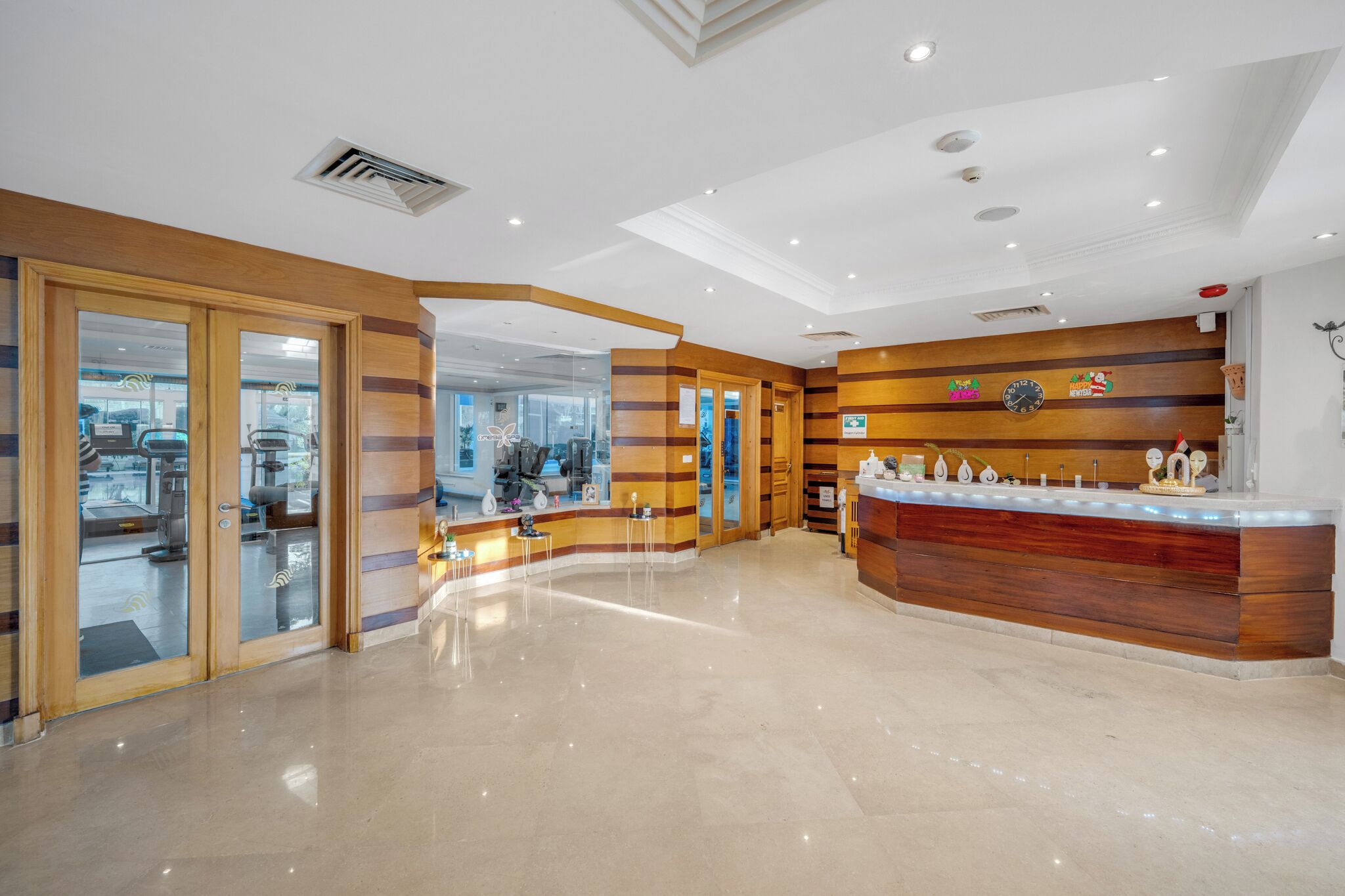 Un vestíbulo de hotel con un mostrador de recepción, puertas de vidrio que dan a un estacionamiento y un reloj en la pared.