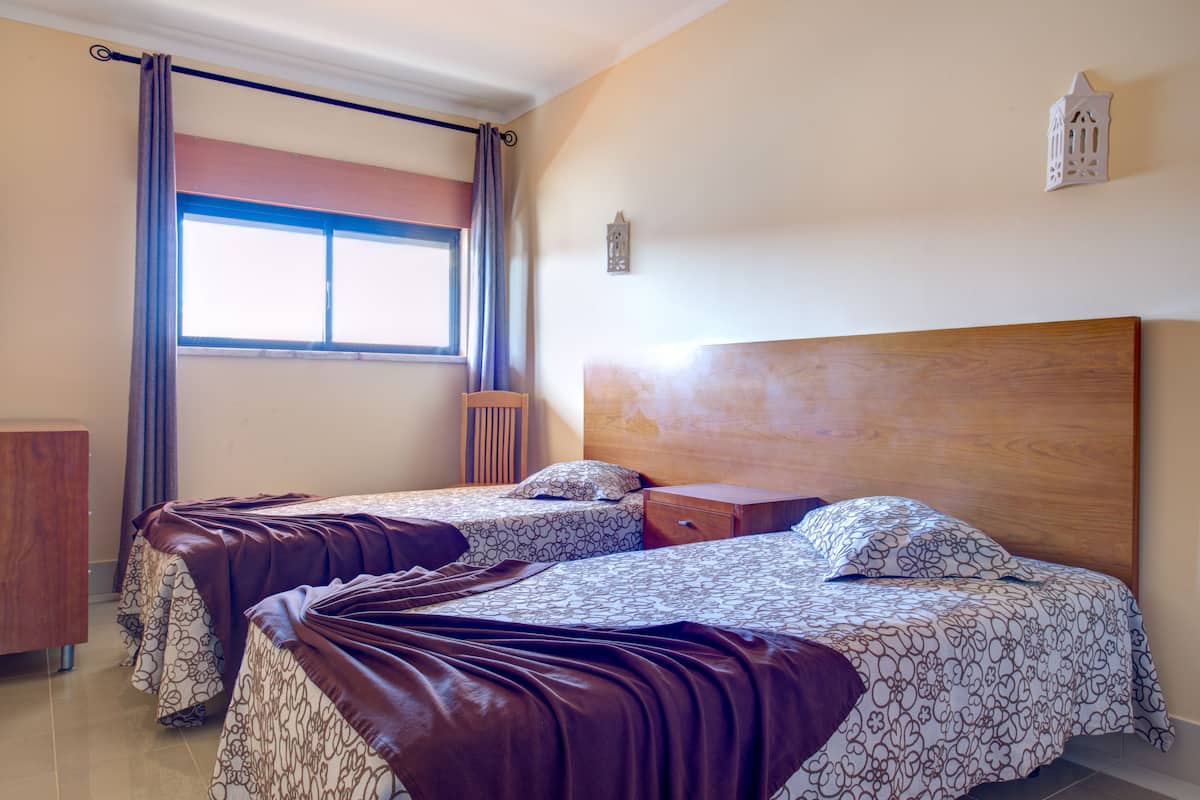 A room with two beds, a window with curtains, a wooden headboard, a nightstand, and a wall-mounted light fixture.