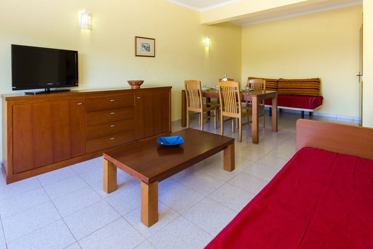 A compact living space with a TV, wooden furniture, a dining table, and a red sofa.