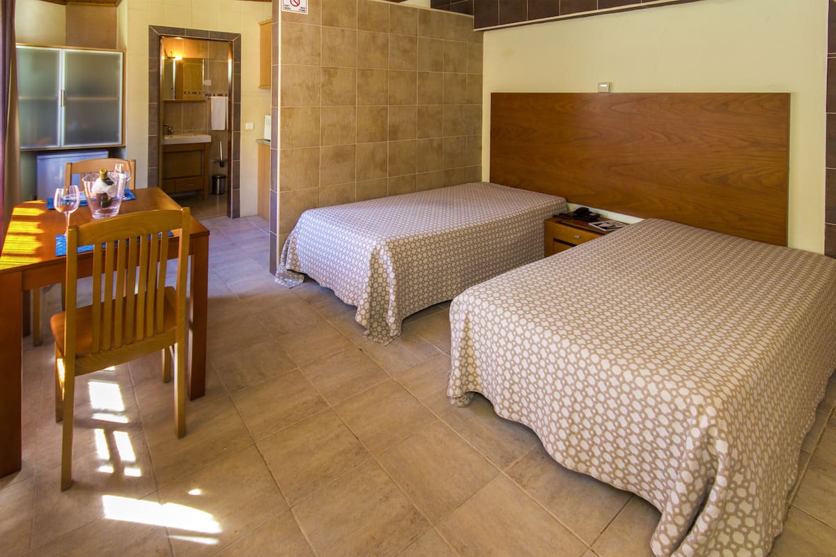A hotel room with two single beds, a wooden desk with chairs, and a small kitchenette area.