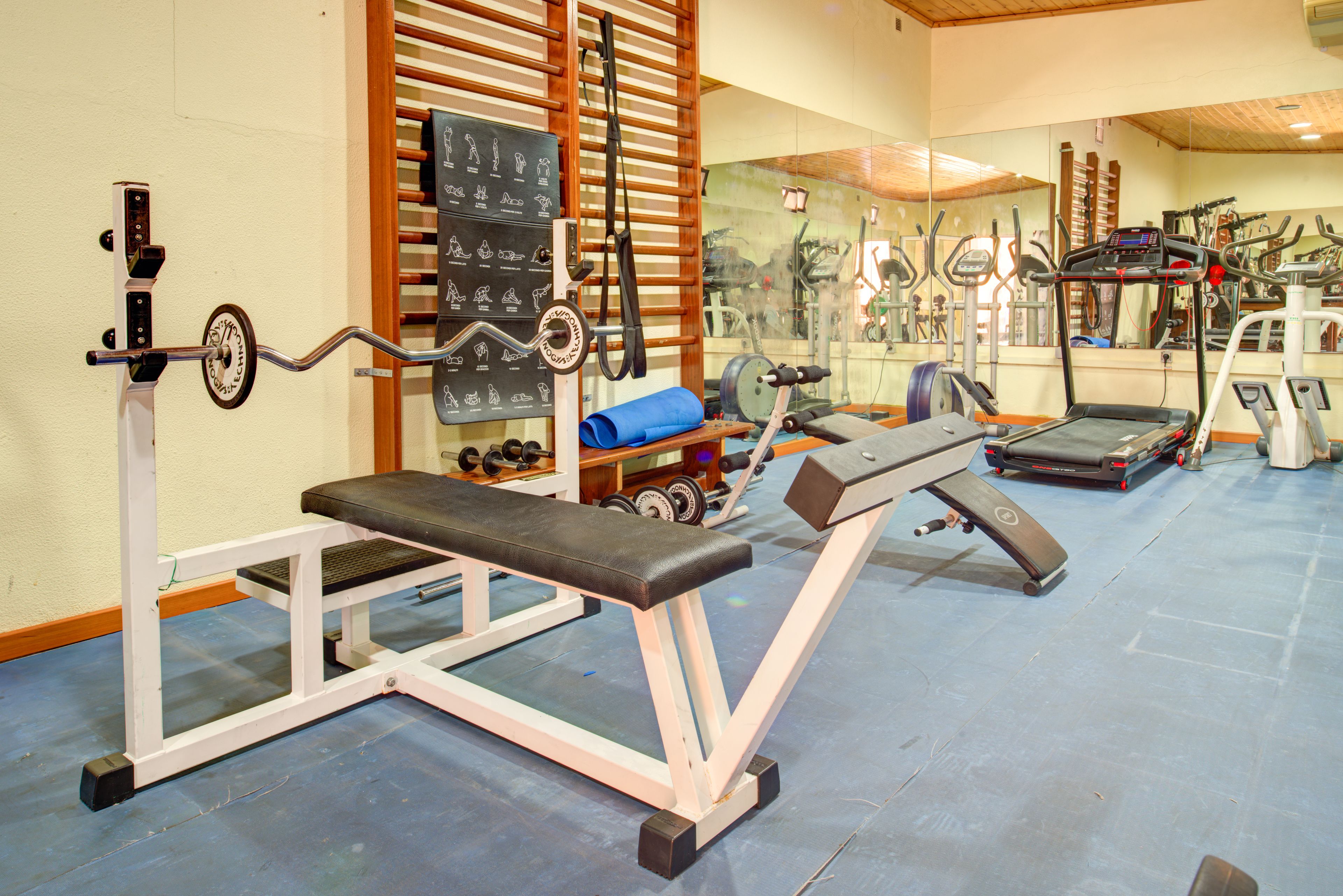 Een sportschool met trainingsapparatuur, waaronder een bank, gewichten en een loopband.