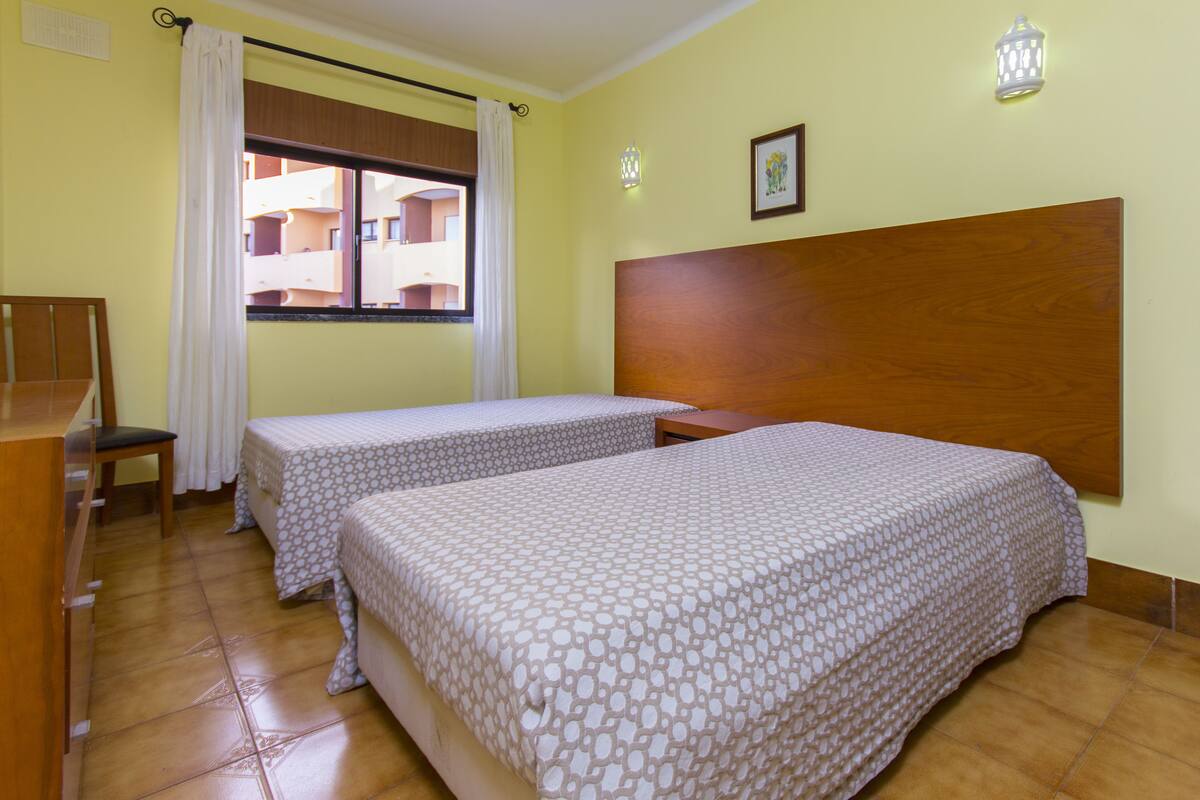 A hotel room with two beds, a wooden headboard, a window with curtains, a chair, and a small table.