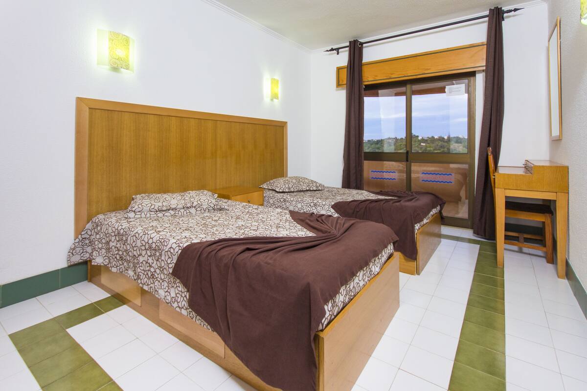 A room with two beds, a wooden headboard, a small table, and a window with curtains.