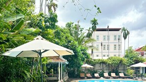 A swimming pool with lounge chairs and umbrellas, surrounded by greenery and a building in the background. - Memoire d'Angkor Boutique Hotel (Siem Reap)
