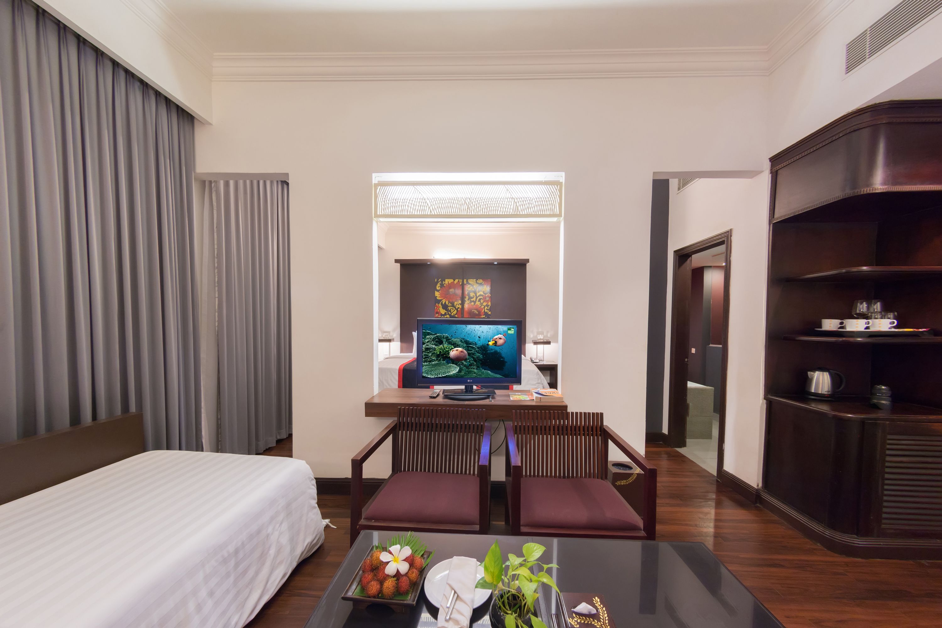 Ein Hotelzimmer mit einem Bett, einem Schreibtisch mit Computer, einem Fernseher und einem Couchtisch mit einer Schale Erdbeeren und einer Vase.